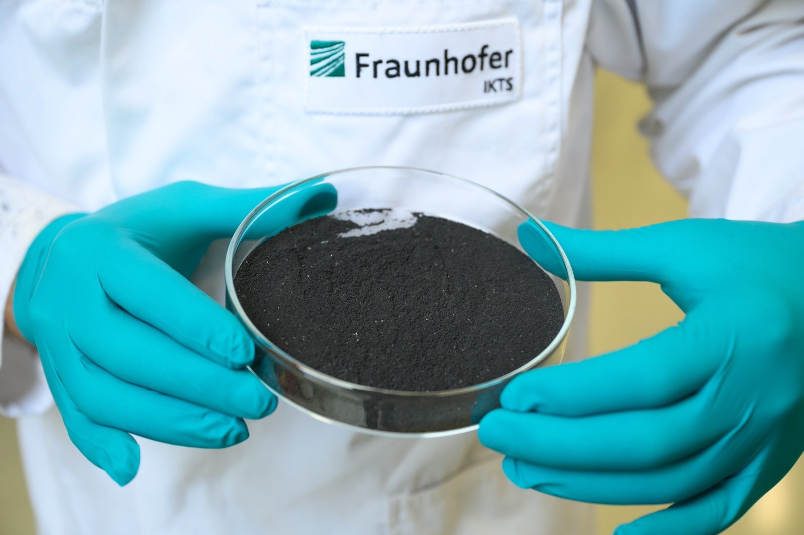 Two hands wearing turquoise latex gloves hold a glass lab dish that contains a dark powder