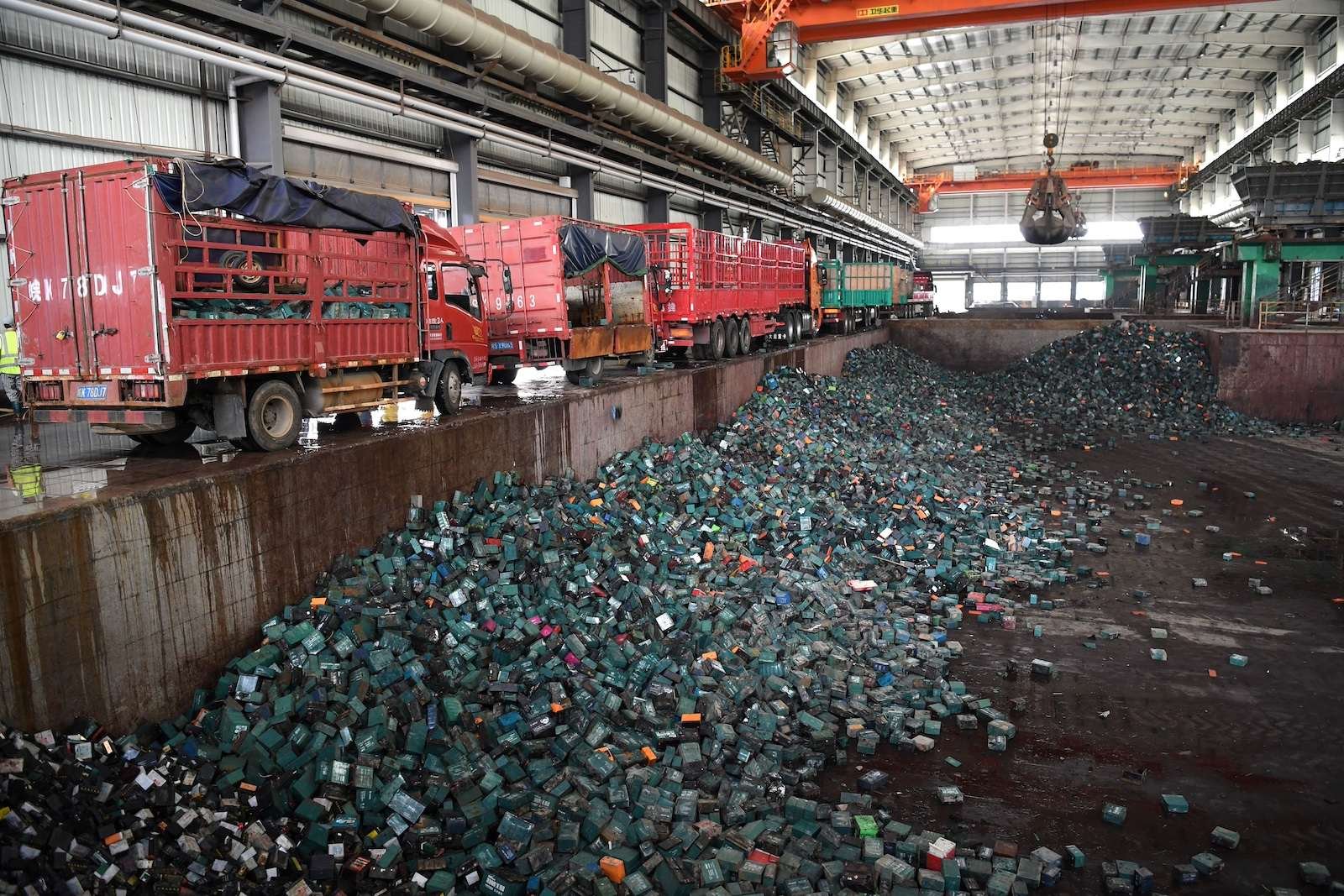 A wide shot of an industrial space where red trucks are parked next to a pit containing huge piles of greenish-gray batteries