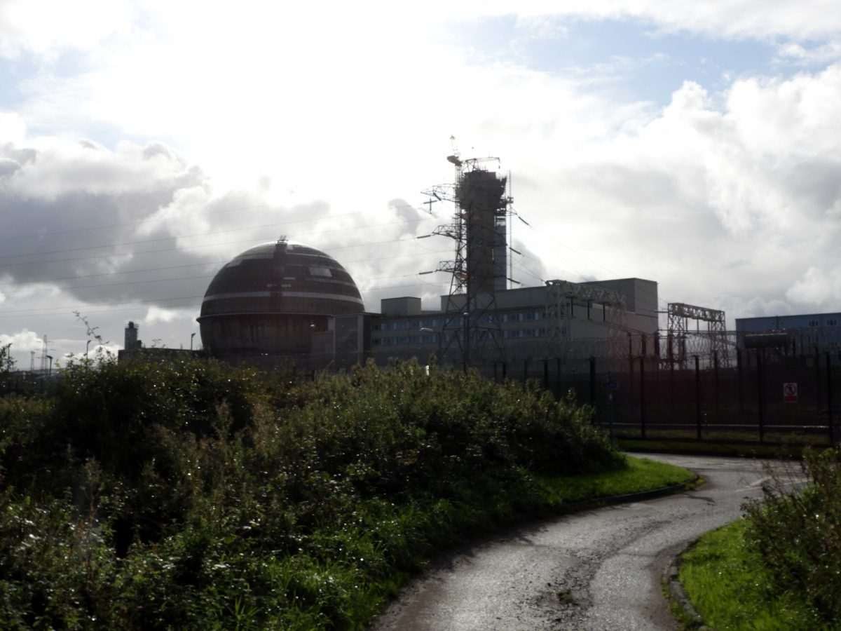 A collection of industrial buildings, chimneys and other structures on a flat, green, coastal expanse of land with a hill or elevated ridge visible in the far distance