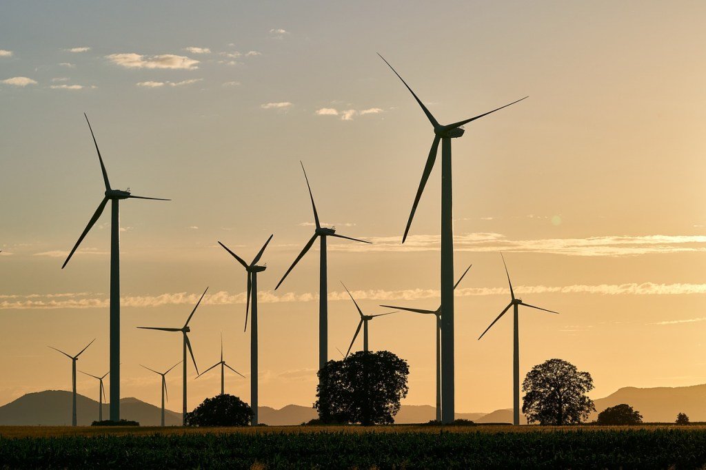 An evening scene with large wind turbines dotted throughout the image over green fields with silhouetted hills in the background.