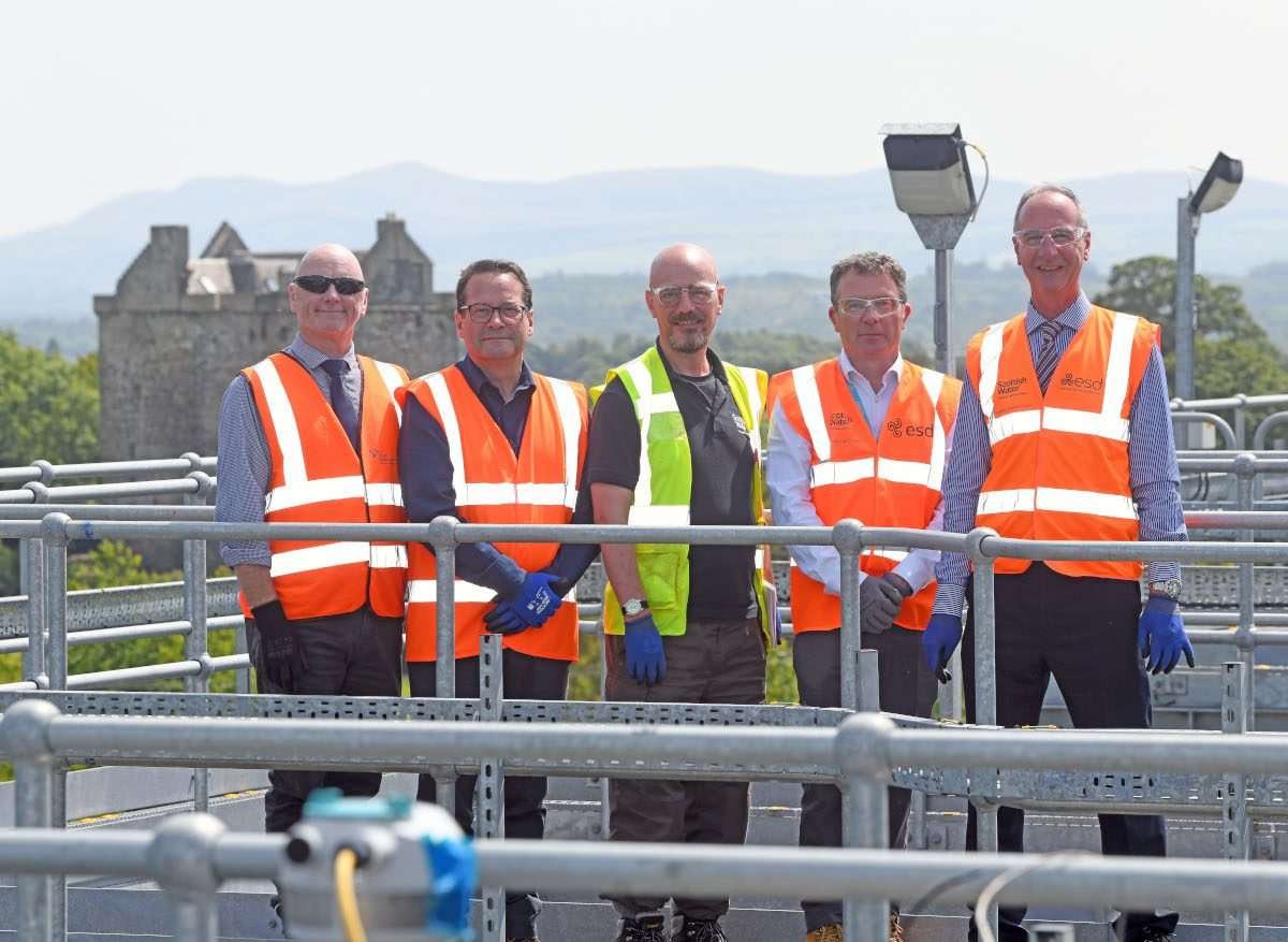 Four men in business attire and high-vis stand abreast each other, facing the camera, in an outdoor semi-industrial location, a wastewater treatment works