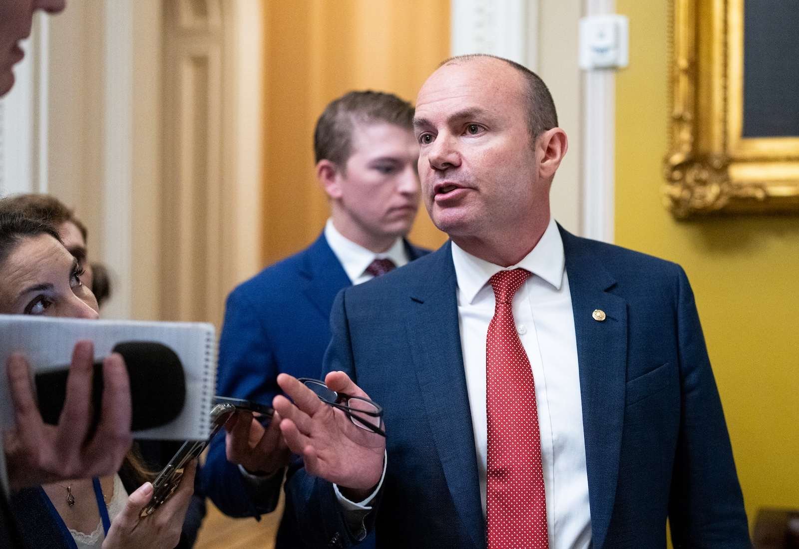 Senator Mike Lee wearing a suit and talking to reporters on his right, inside a room with yellow walls