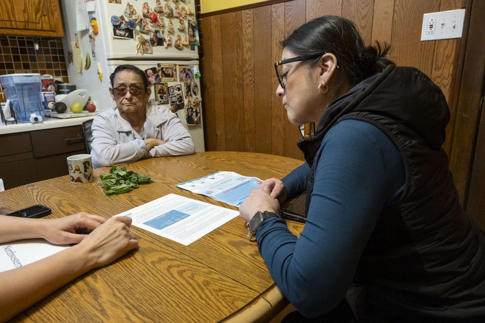 two women sit at a kitchen table looking over flyers with charts and information