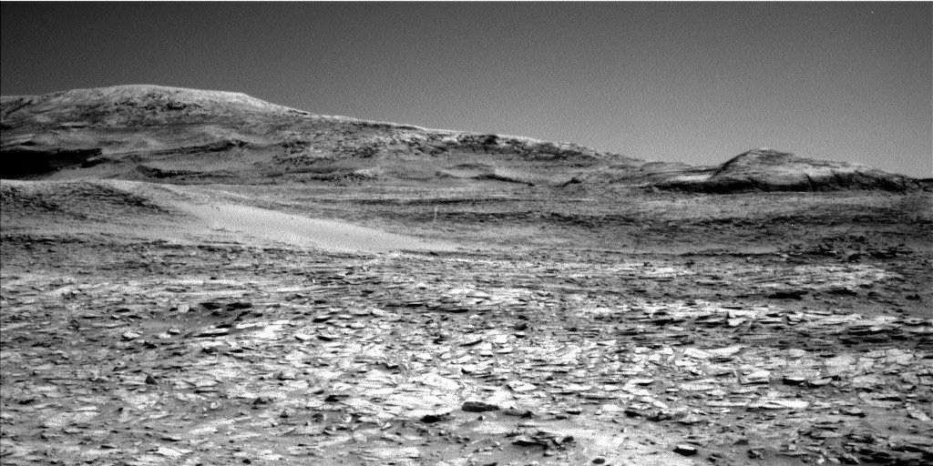 A grayscale photo from the Martian surface shows rocky ground from the front of the image to a horizon line where a series of gently sloped hills rise up.