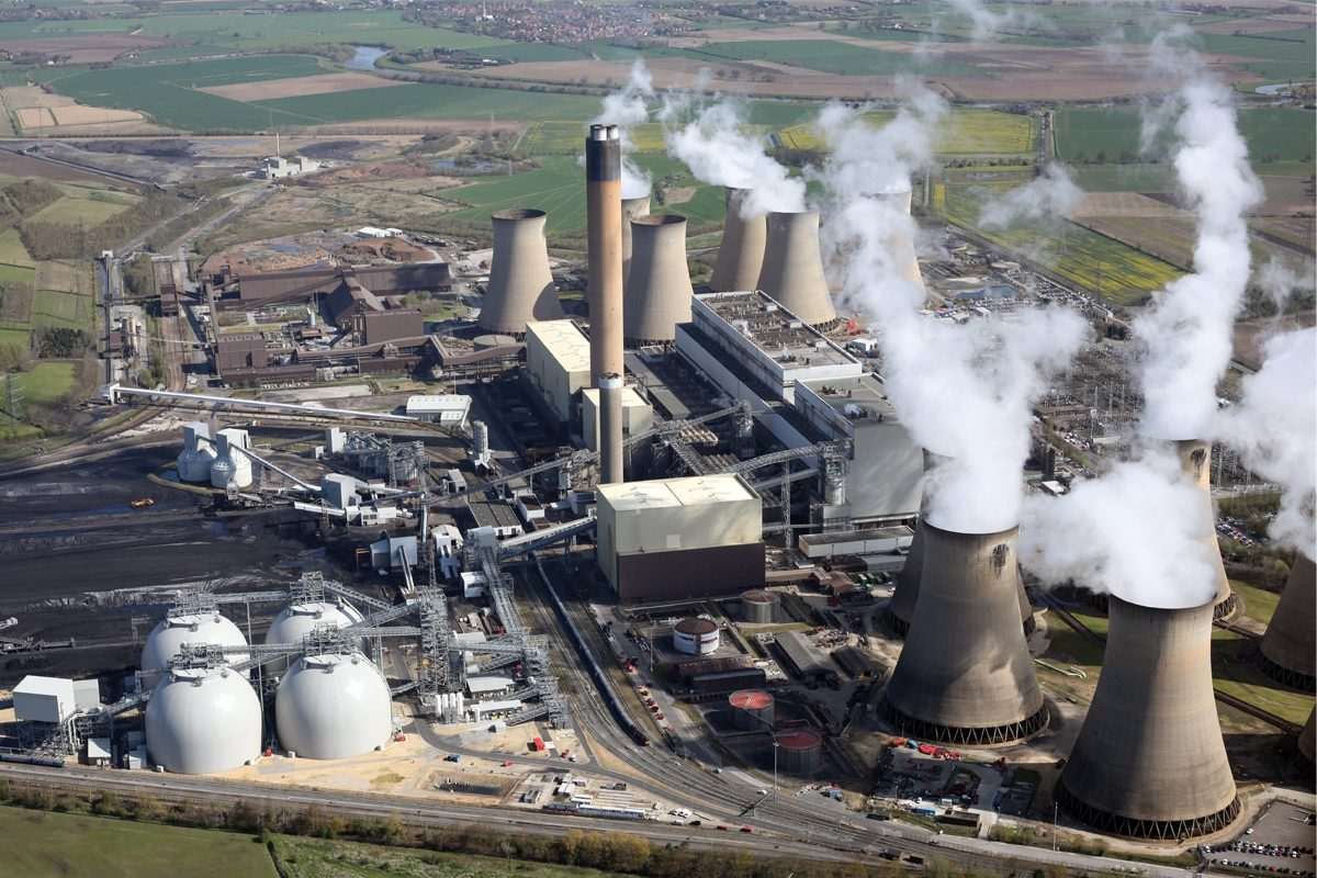 Aerial view of Drax power plant showing a multitude of cuboidal buildings and several large, multi-storey chimneys from which opaque white fumes are visibly drifting, all set within a green rural landscape