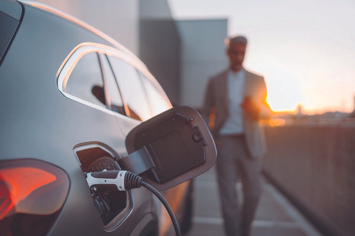 Close up of charging inlet of modern electric vehicle, plugged into a charging point, as a man in business atire walks towards the camera, and the whole scene takes place outdoors against sunrise or red-hued sky in an urban location