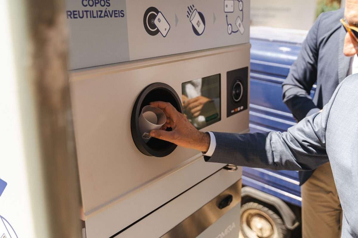 A man deposits an empty coffee cup in a circular slot on an oblong kiosk in an outdoor, bright location