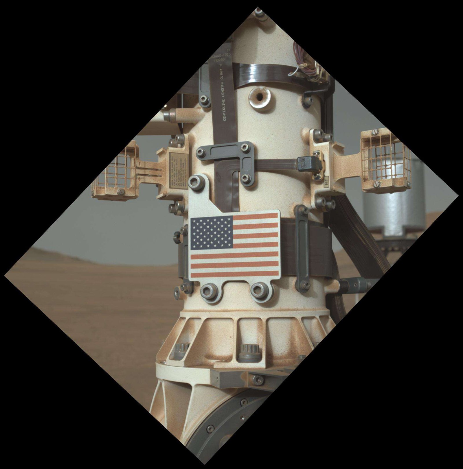 A small metal American flag attached to the Mars Perceverance rover's mast. The mast is white with silver metal pieces attached to it. There is red dust all over the mast. The image appears in a vignette that looks like a rectangle rotated diagonally; the rest of the image is dark.