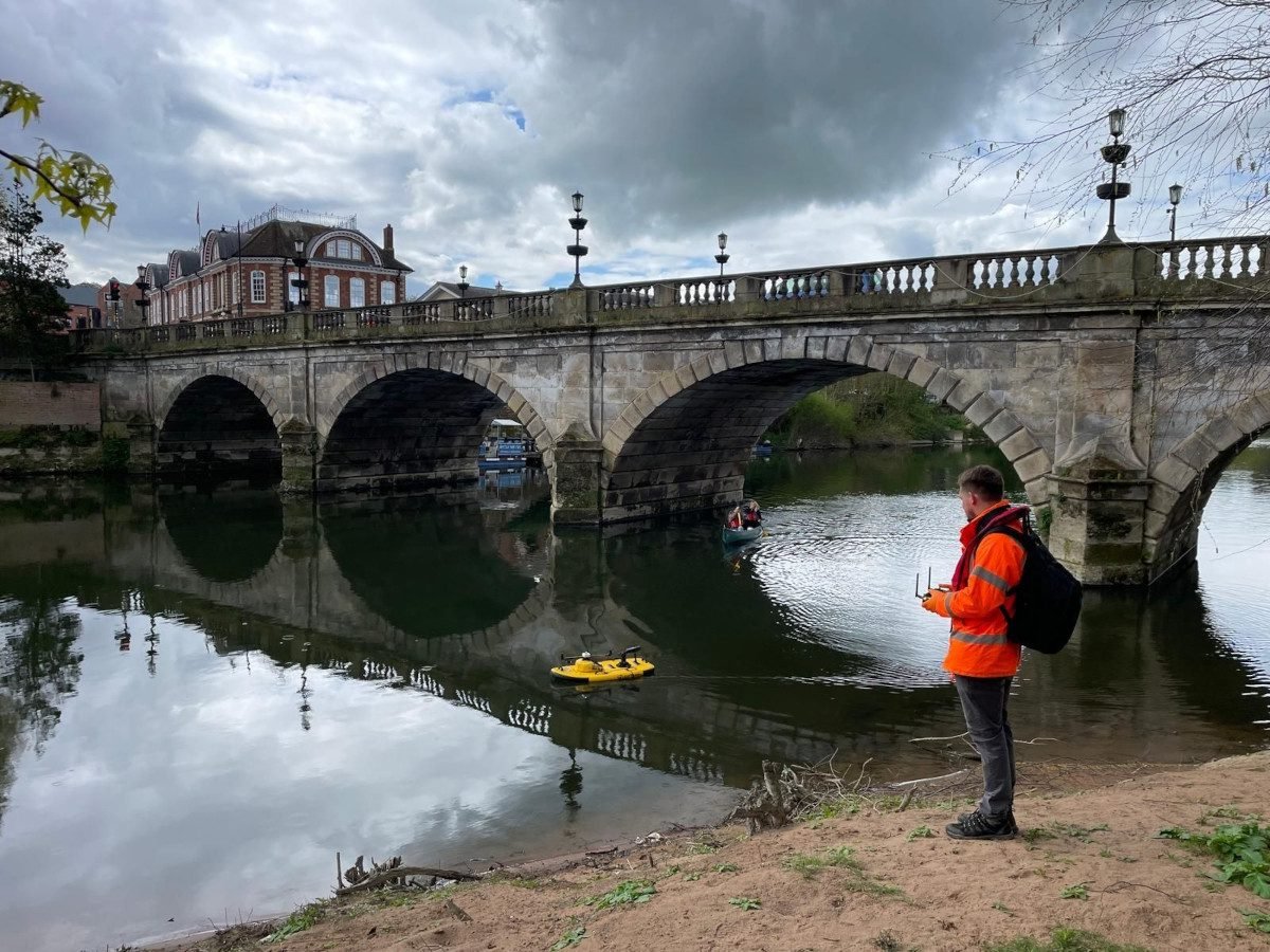 A man stands on the bank of a river, operating a remote control pad, while miniature autonomous vehicle is visible out in the water, and in the background an old-fashioned stone road bridge crosses the river, with buildings visible on beyond the bridge, on the other side of the river