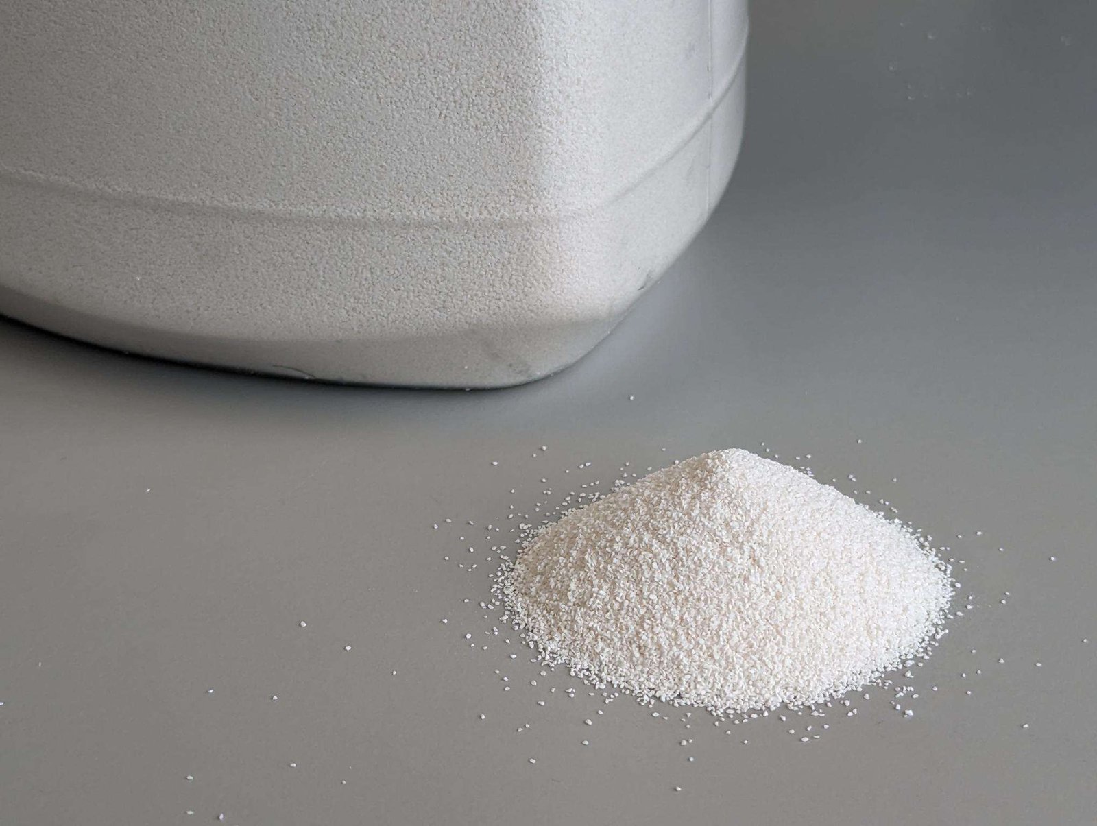 A fine white powder is piled up in a cone shape on a flat surface with white plastic object nearby that might be the edge of a plastic container