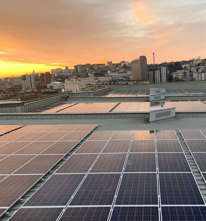 Rooftop solar panels on flat roof, and themselves lying flat, while background of image reveals UK urban cityscape seen at roof level, beneath orange-hued sky