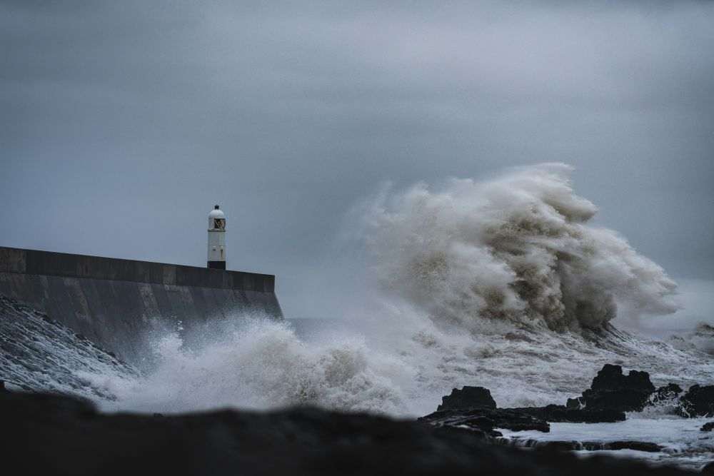 A modern stone jetty is struck by waves during a storm event, with dark rocks visible below, set against background of grey and white water and sky