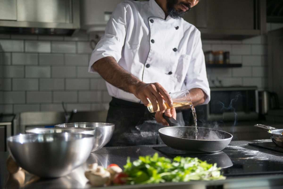 Chef in restaurant kitchen or similar setting pours transparent fluid from a glass container into a frying pan