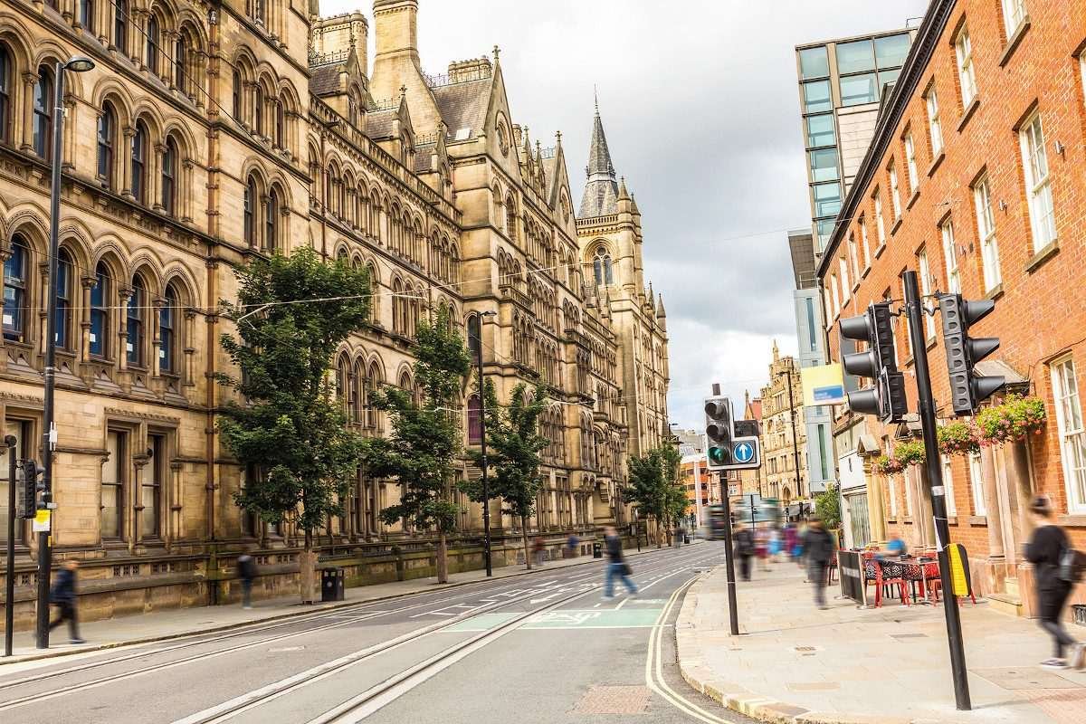 Image of a main street in Manchester with tall red-brick Victorian structures on either side and a few people visible walking through the scene
