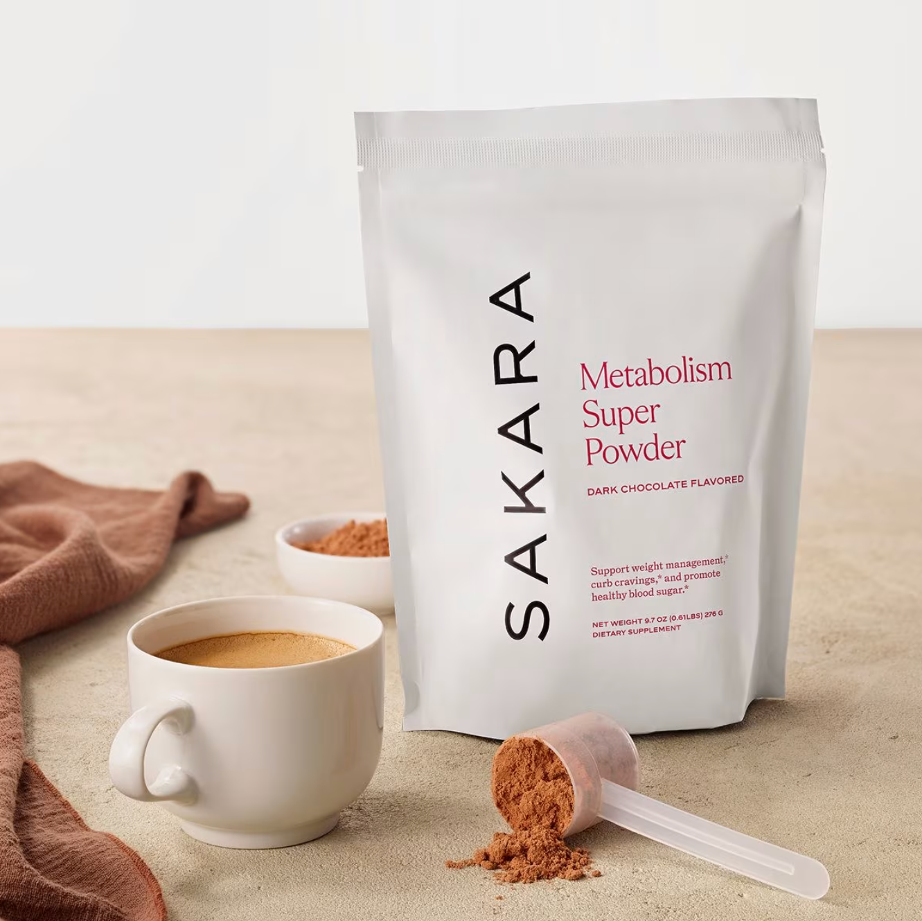 A bag of Sakara Metabolism Super Powder stands upright next to a white mug of prepared drink, a scoop of powder, and a brown cloth on a beige surface.