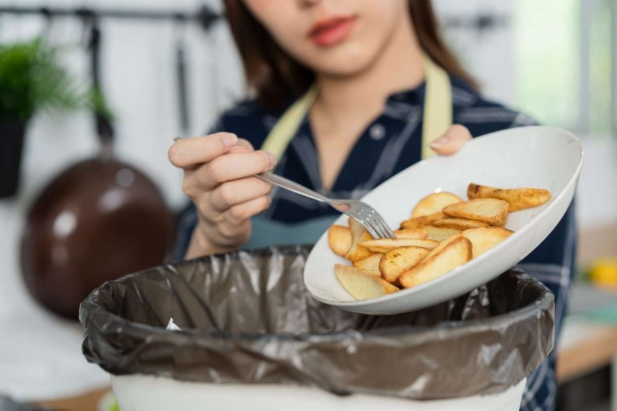 A plate of frites, or chips, is being tilted to empty contents into a waste bin, which is being held by a pair of hands belonging to an oriental woman, seemingly a restaurant worker, the lower portion of whose face is visible