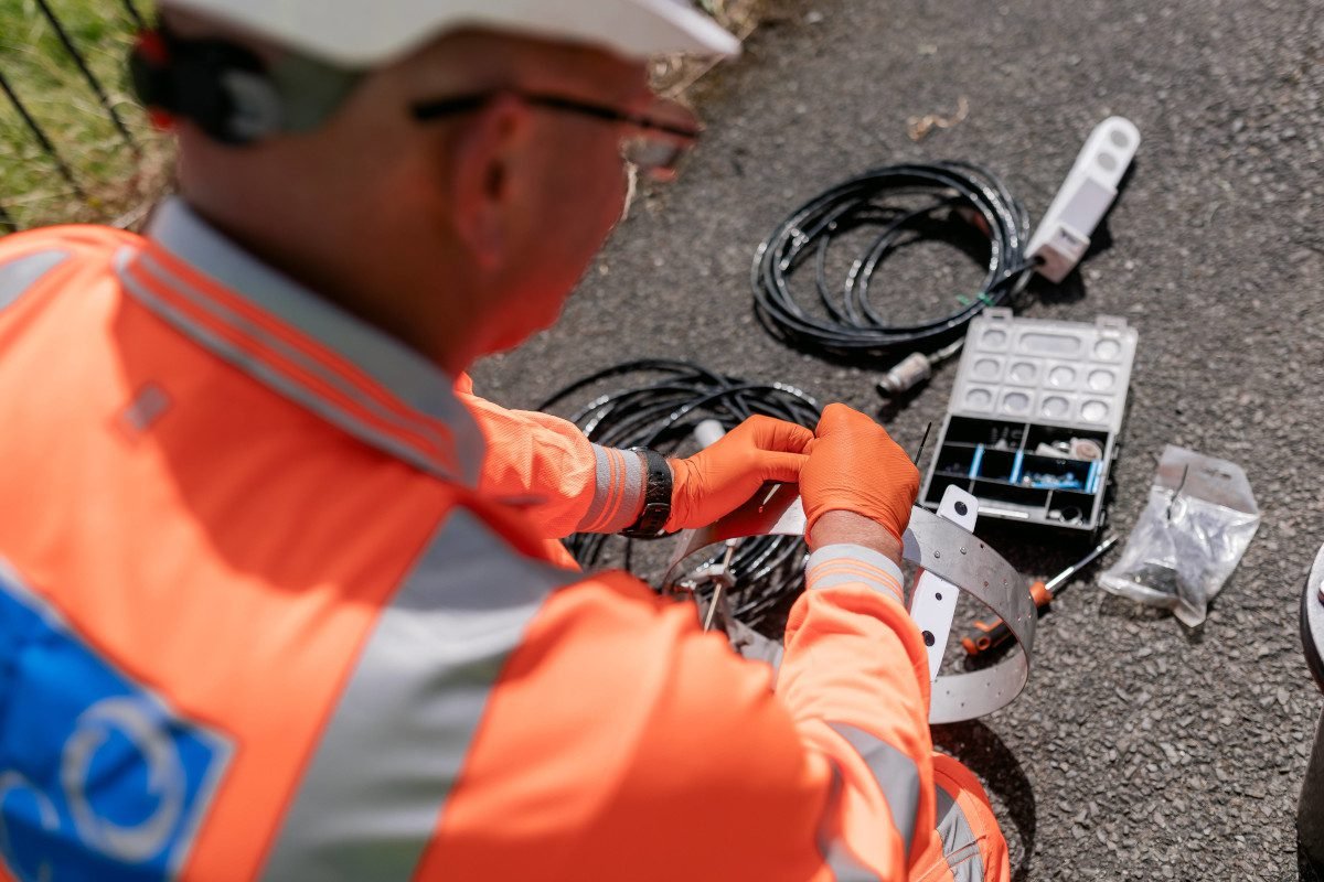 Technician in orange overalls and hard hat crouches to work on electrical equipment and wires spread out on the ground beneath him, in an outdoor location like a river bank