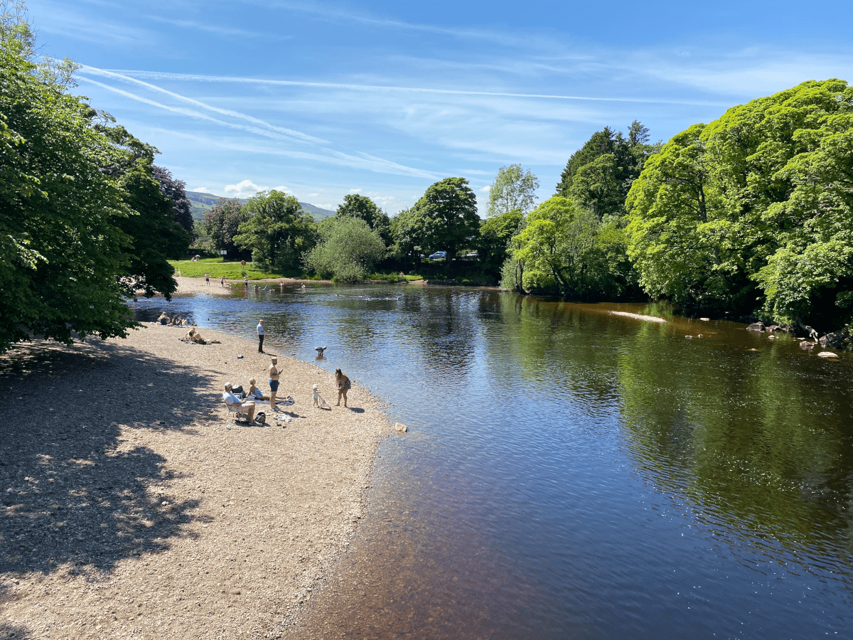 Slightly elevated view of bending section of the river Ilkley, 10 or 20 metres wide, with trees on either side, as if in a rural location, and one bank comprises a flat, large beach area of sandy-coloured stones with families and people sunbathing and reclining in the sun