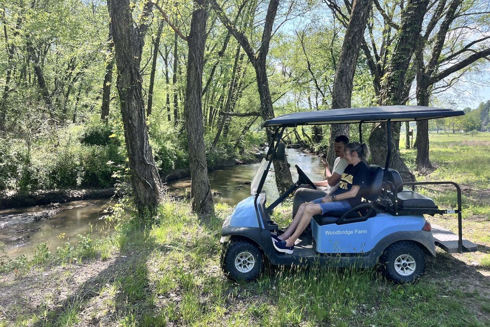 a man and a woman in a golf cart drive to the edge of a woodland river