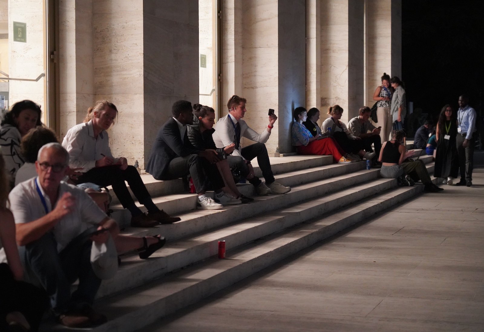 People sit on the steps of a large building at night, backlit by interior lighting