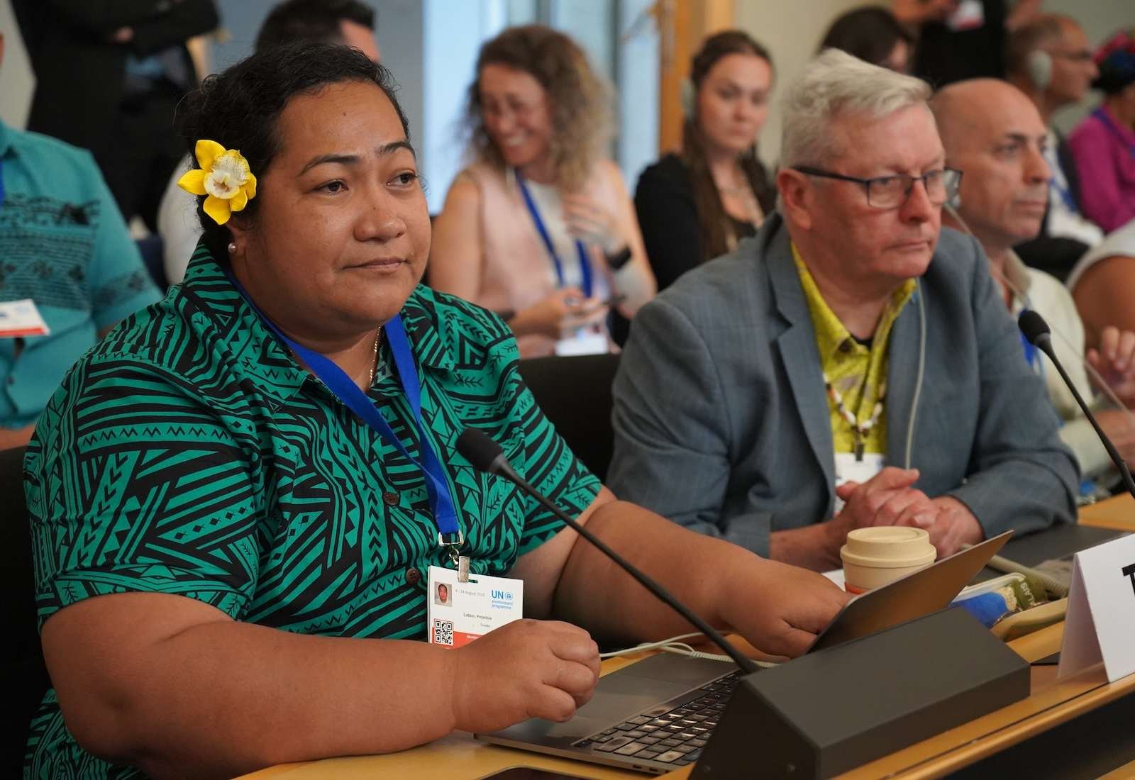 A woman in green with a flower in her hair sits at a table with a microphone in front of her. A man is to her left, seated.
