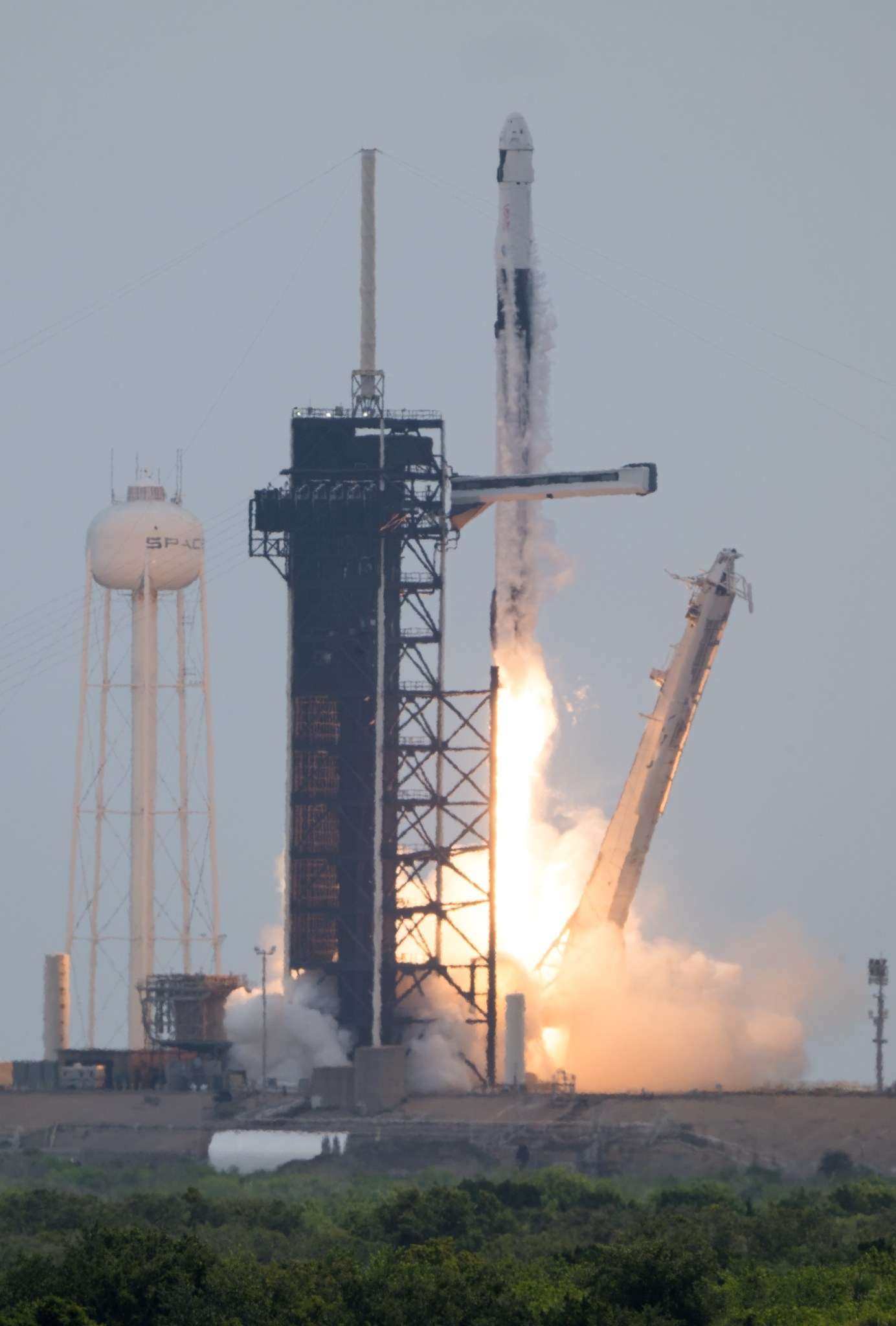 A rocket lifts off from the launchpad, leaving a trail of flames and plumes of vapor in its wake. A white structure and the black gantry can be seen on the left. On the right, a white arm falls away. The photo is taken from a distance.
