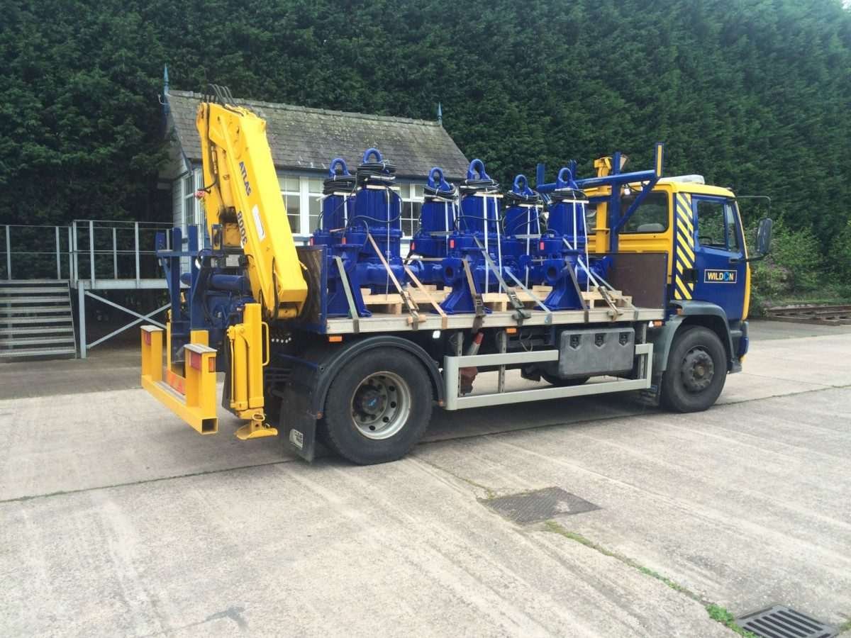 Flatbed lorry situated on a road, and the cargo platform is loaded with a number of upright, long, semi-cuboidal structures, which are presumably pumps