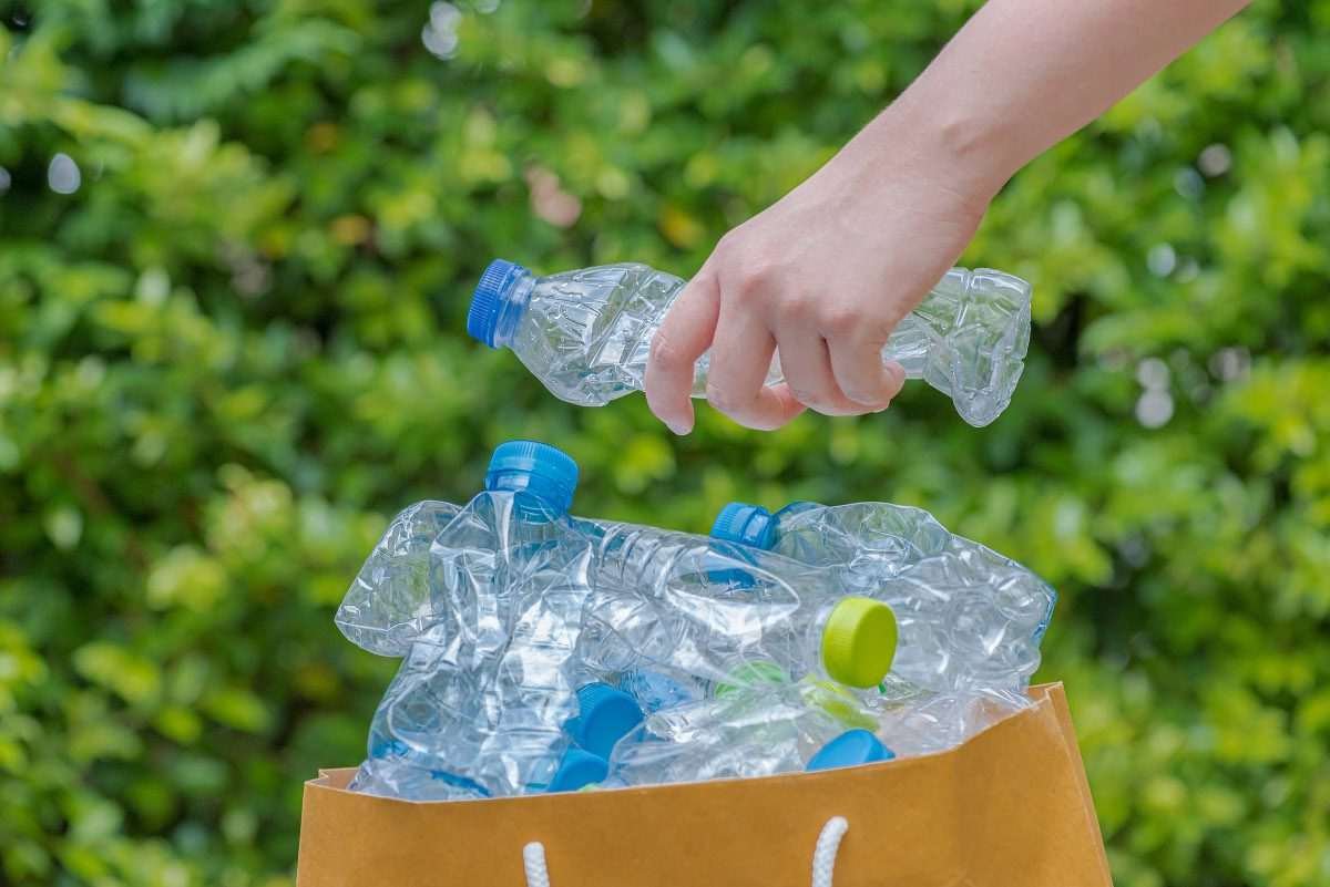 A hand is clenched around a plastic drinks bottle as it is deposited in a paper bag full of similar bottles