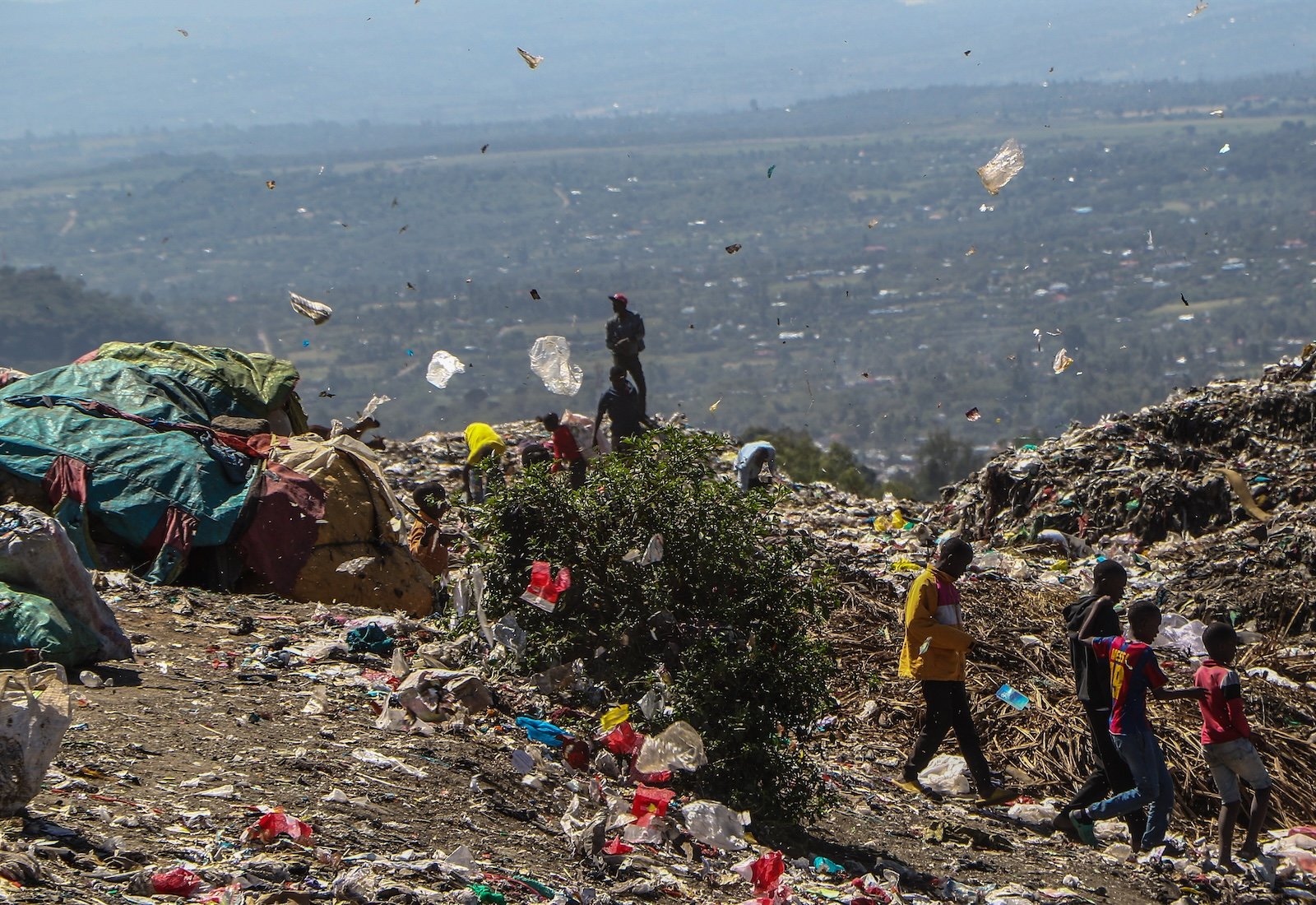 View of a large dump site with plastic bags blowing in the wind and waste pickers walking on top of piles of garbage.