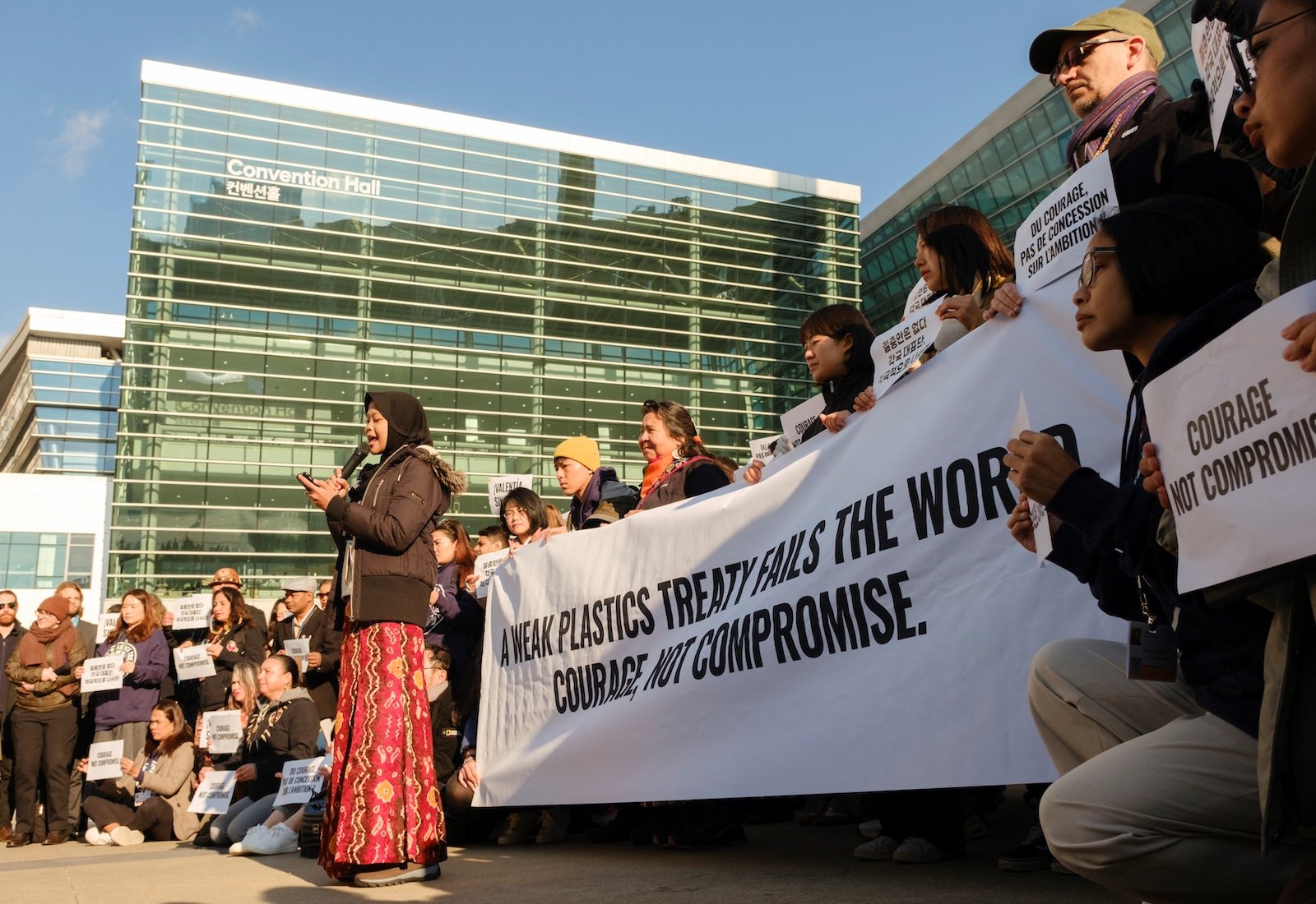 Side view of a protestor reading a speech. Behind her, people hold up a banner reading, "A weak plastics treaty fails the world. Courage, not compromise."