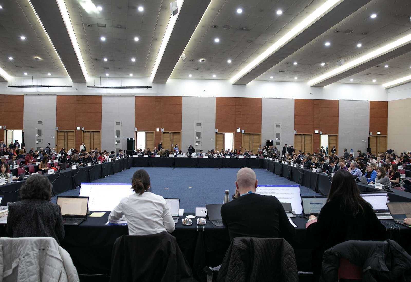 Delegates sit at tables arranged in a very large circle, in a brightly lit room.