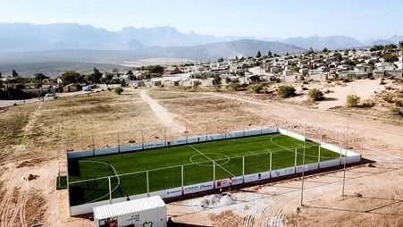 A small football pitch of artificial green turf, surrounded a fenced enclosure situated within arid surroundings, possibly an unpopulated area, with mountains visible in the distant haze
