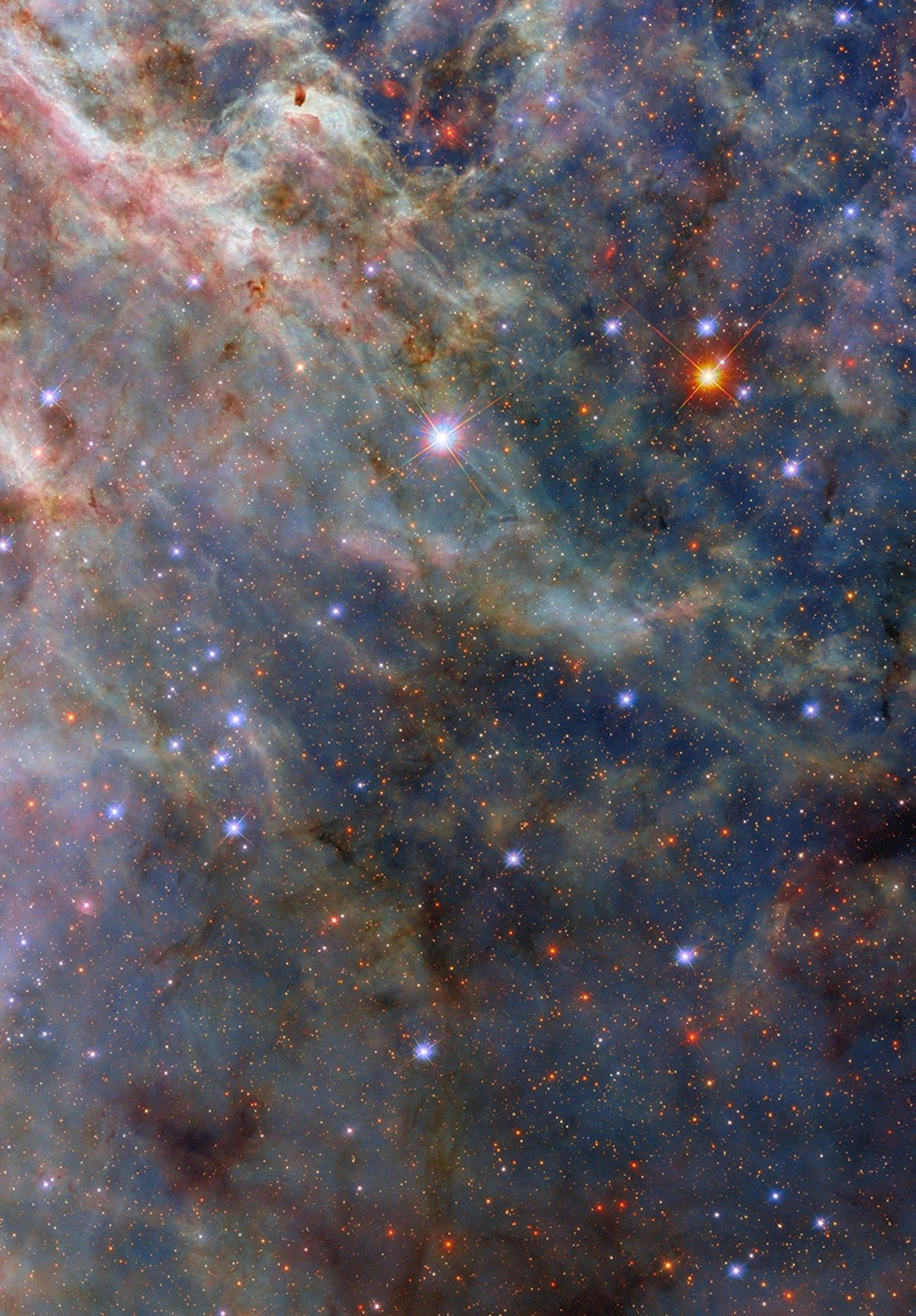 A colorful nebula. The image’s top-left is filled with layers of fluffy pink and greenish clouds. Long strands of green clouds stretch out from here; a faint layer of translucent blue dust combines with them to create a three-dimensional scene. A sparse network of dark dust clouds in the foreground adds reddish-black patches atop the nebula. Blue-white and orange stars, from our galaxy and beyond, are spread throughout the clouds.