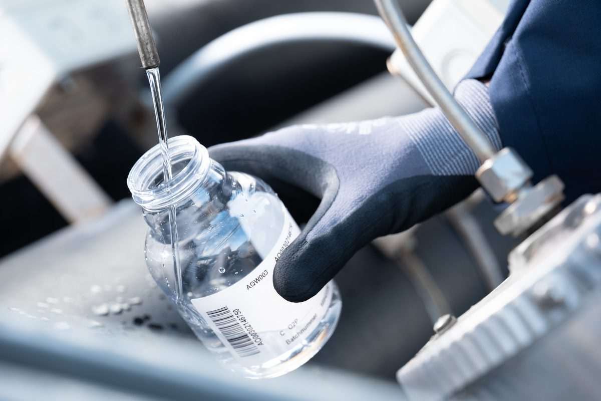Gloved hand holds glass container receiving jet of water from a small tap, with all elements indicating a scientific laboratory or similar environment