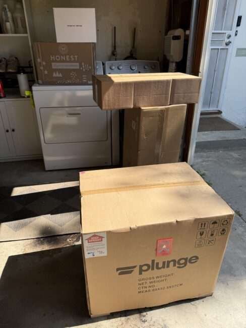 Several cardboard boxes of various sizes, including ones labeled "plunge" and "Honest," are stacked in a laundry area near a washer and dryer.