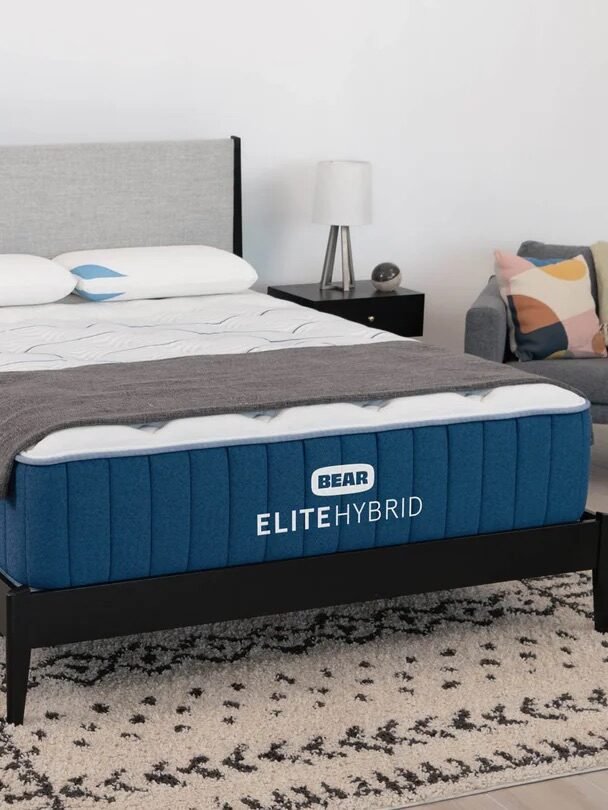 A modern bedroom featuring a Bear Elite Hybrid mattress on a black bed frame, two pillows, a gray throw blanket, and an armchair; a bedding award badge is displayed in the foreground.