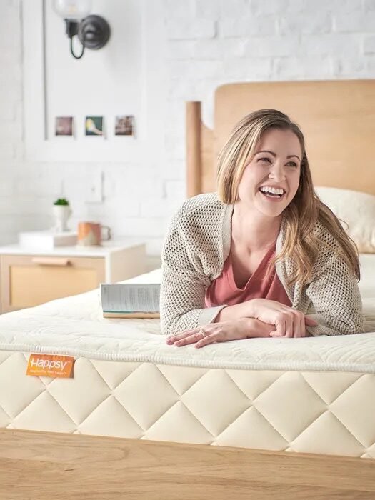 A woman lies on a bed, smiling and holding an open book. The mattress has a quilted pattern and a visible Happsy label. The room has a white brick wall and wooden furniture.