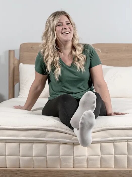 A woman with long blonde hair sits on a bed, smiling, with legs extended and feet pointed toward the camera.
