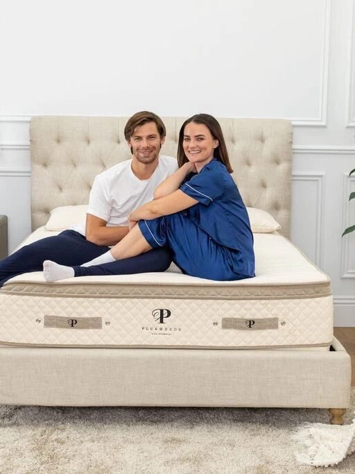 A man and woman sit together on a bed in a bright, neatly decorated bedroom with plants, a nightstand, and a lamp.