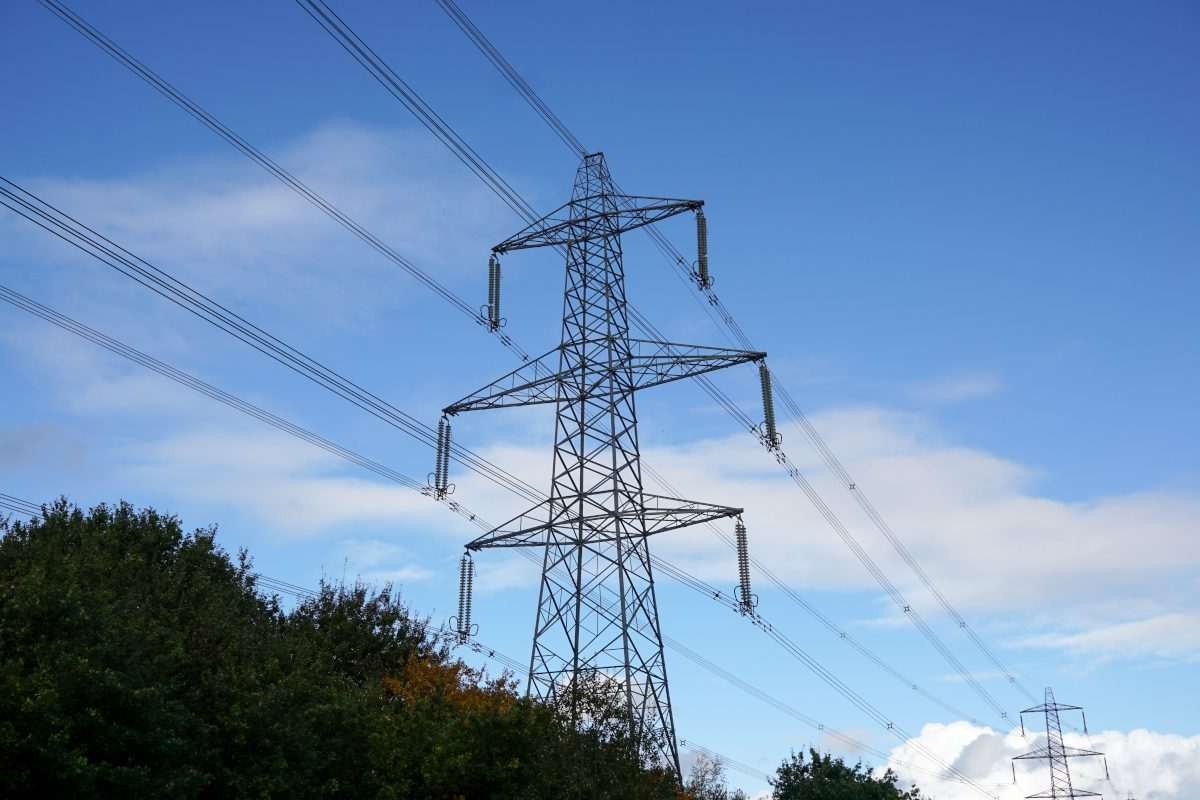 Overhead electricity pylon set against a blue sky with scattered clouds and with the protruding tops of trees or foliage just visible near the bottom of the frame