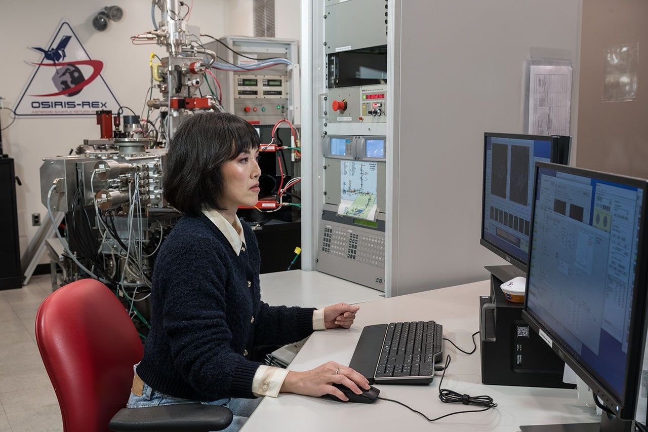 A picture of a woman sitting at a desk, working at a computer. She is in a lab, and a large, complex machine can be seen behind her. On the wall is a plaque that has the logo for the OSIRIS-REx mission.