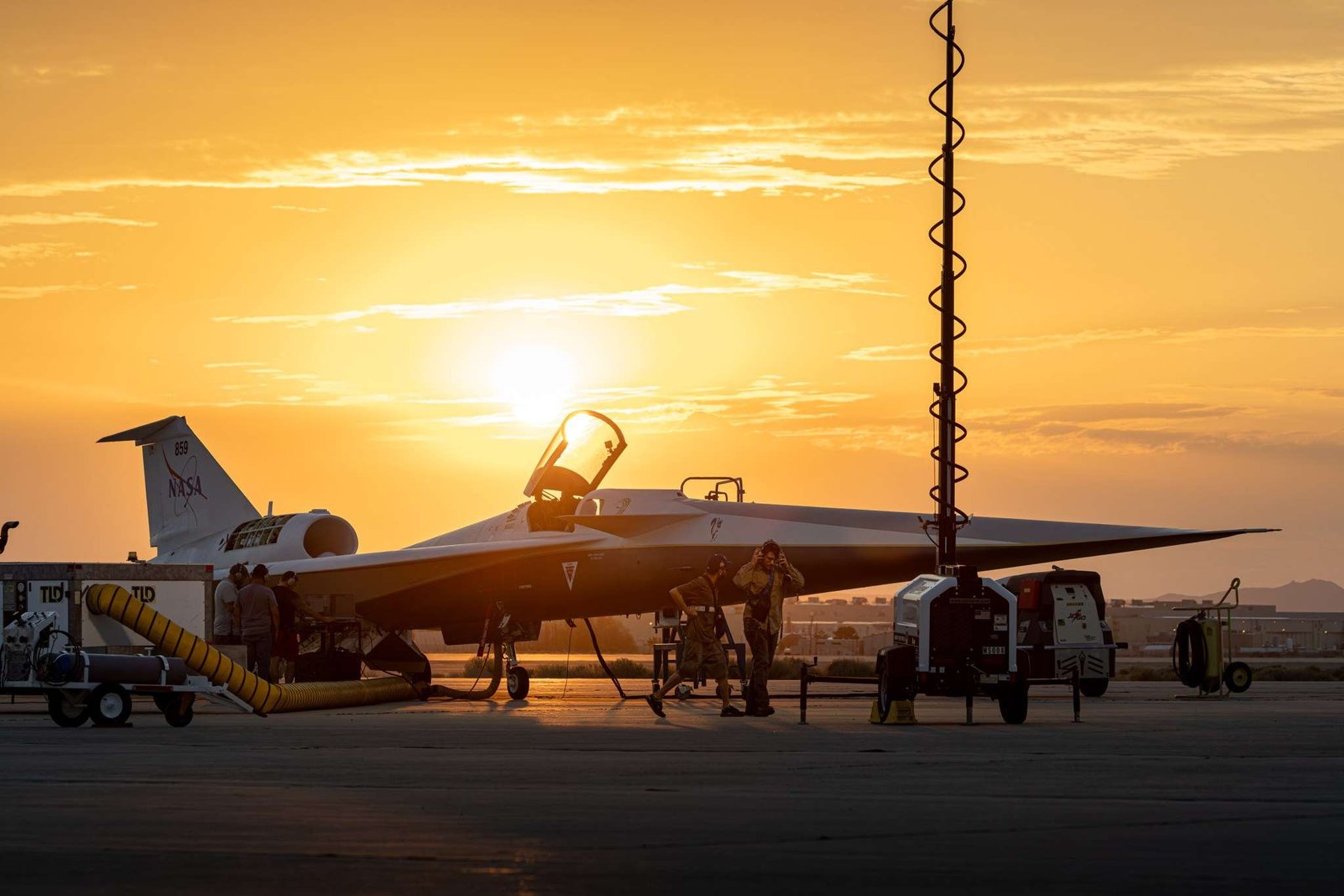 NASA’s X-59 quiet supersonic research aircraft is seen on the ramp at sunrise with its canopy open. Orange and gold light filters through the clouds behind the aircraft, highlighting the aircraft’s shape. Several ground crew members are visible around the aircraft, along with support carts. A vertical light pole with a coiled cable can be seen in the foreground.