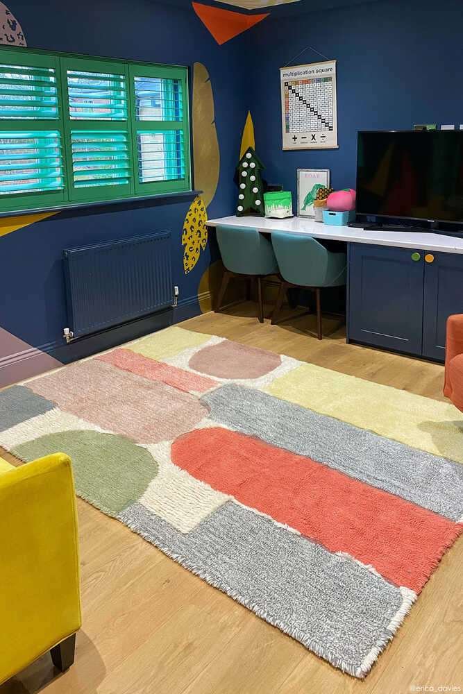 A colorful modern room with a geometric patterned rug, green window shutters, a TV on a blue cabinet, and a desk with two chairs against a dark blue wall.