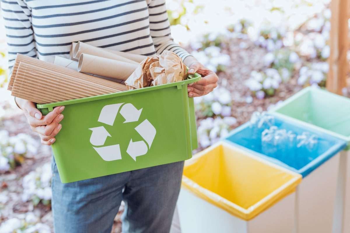 Person carrying plastic cuboidal box with a recycling logo on it, while recycling bins are visible in the background