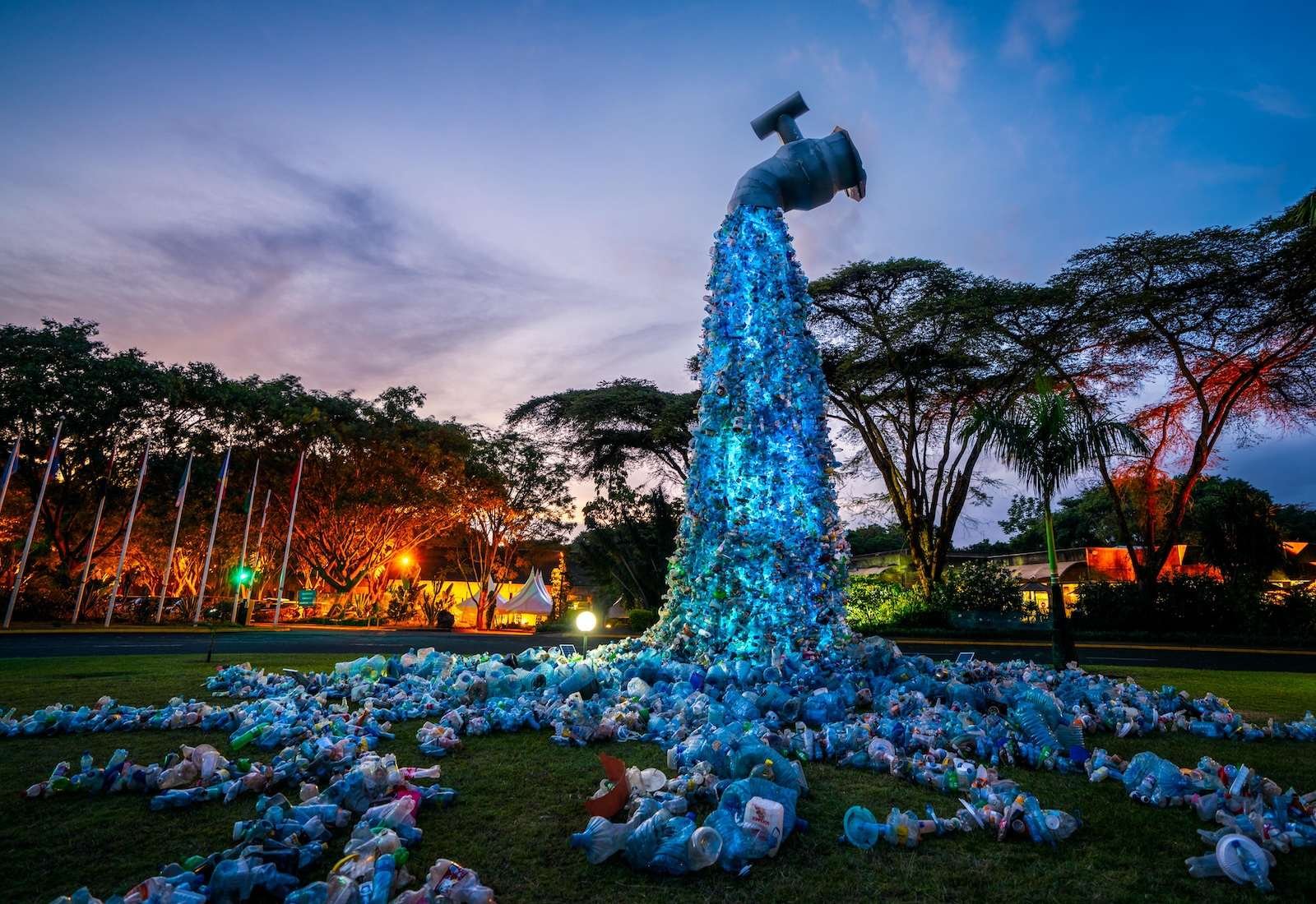 Sculpture of a water fountain at night, with plastic trash flowing out of it onto grass. Trees in background.