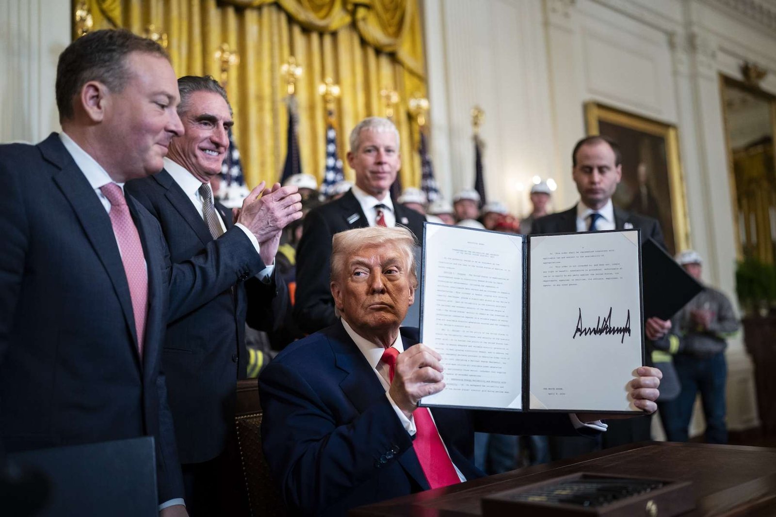 President Trump sits at a table holding up a signed executive order while cabinet members look on. Behind them is a gold curtain.