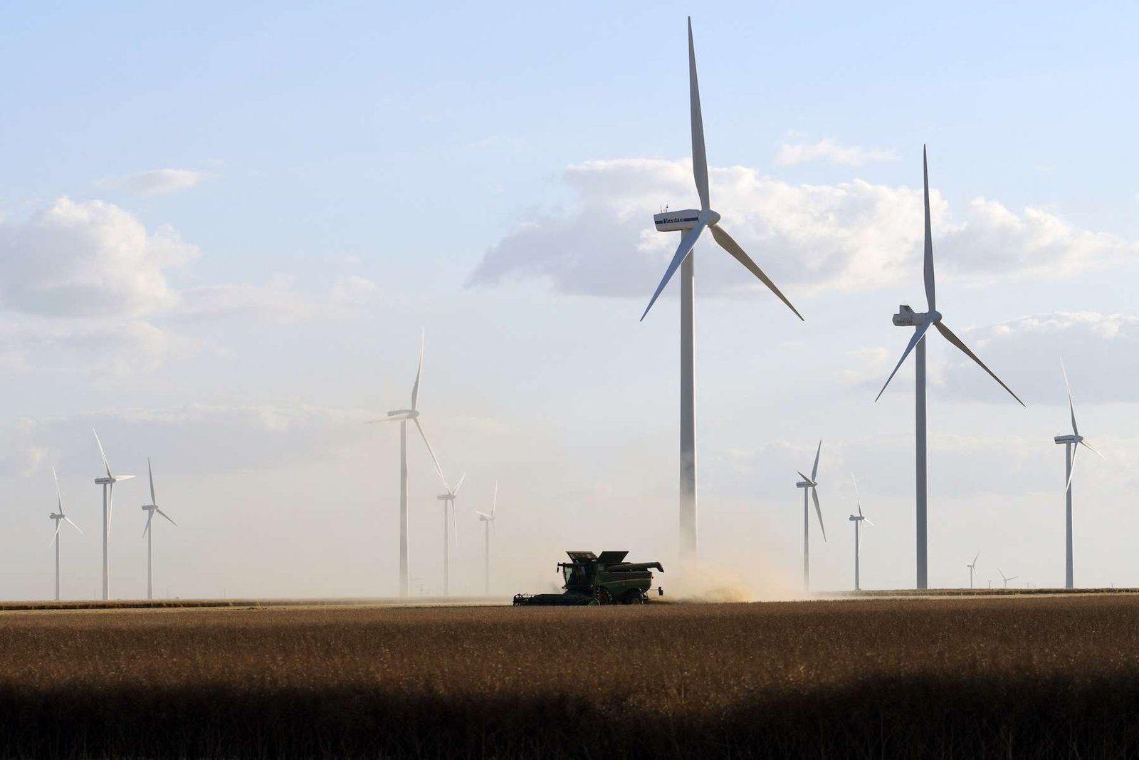 Wind turbines in a wheat field. The sky behind them is blue, but a thresher throws up wheat, making it look like dusk is approaching.