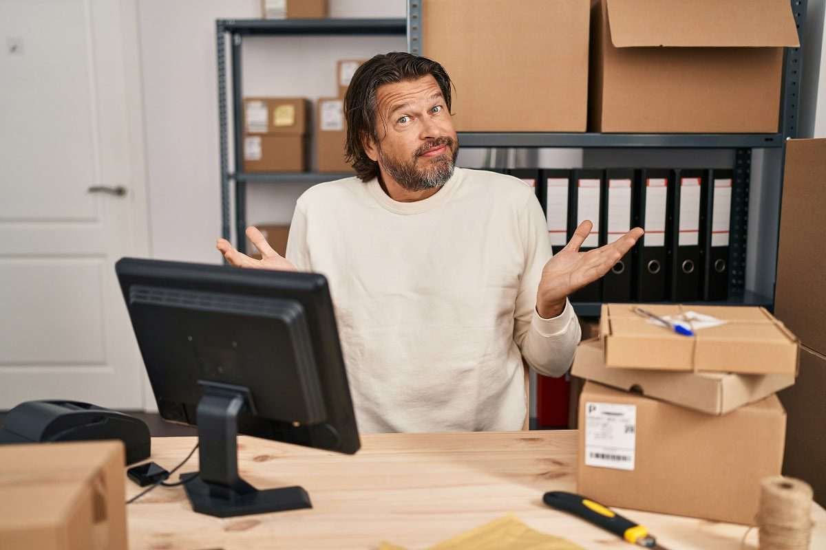 A middle-aged man sitting behind a desk in semi-casual attire, in a very basic office space, with a computer screen in front of him, with facial expression and gesture of two hands raised seeming to say