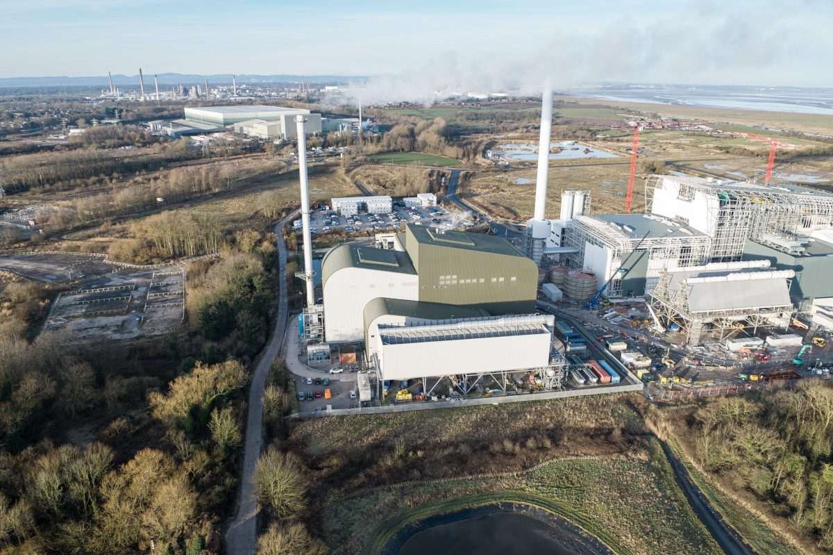 Aerial view showing large cuboidal, partly-curved structures - some kind of industrial facility - with several prominent chimneys, all situated within flat, mostly rural landscape with visibility extending to the horizon and one or two other large industrial and chimney-strewn sites visible in the far distance