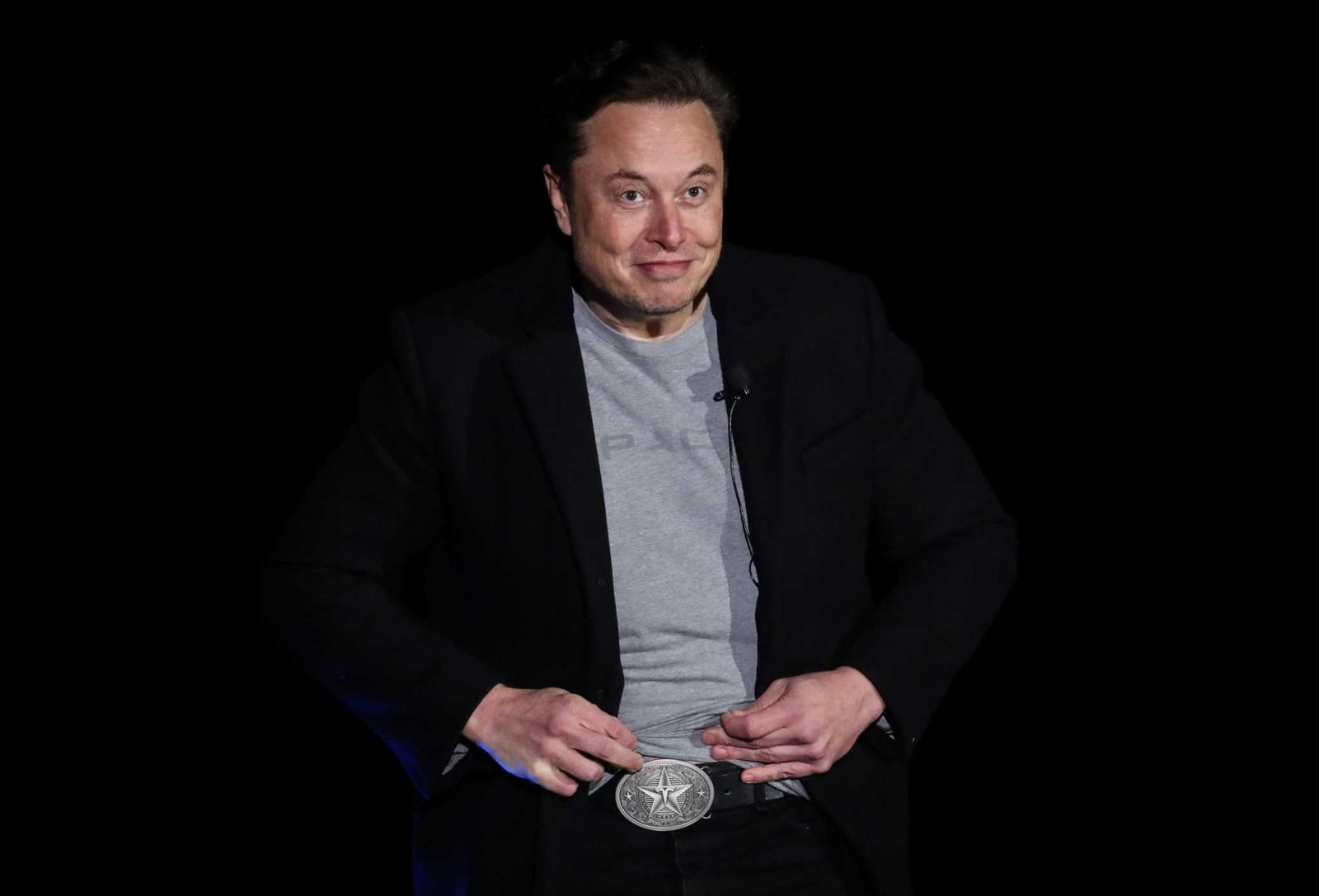 A man in a gray tee shirt and black blazer points to a large silver belt buckle